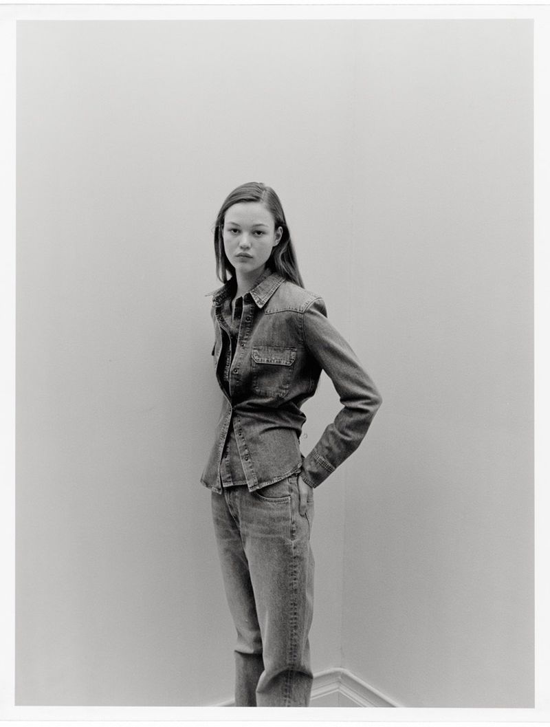 Young woman in denim shirt and jeans stands against a light wall in a neutral pose, captured in a studio-like setting with natural lighting.