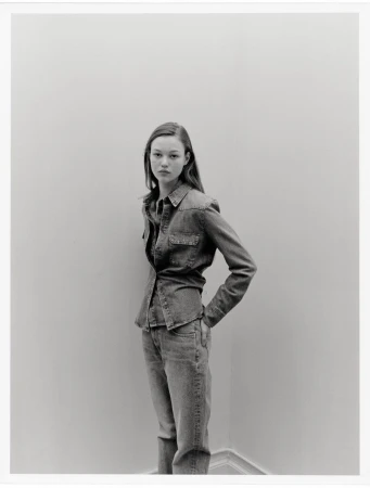 Young woman in denim shirt and jeans stands against a light wall in a neutral pose, captured in a studio-like setting with natural lighting.