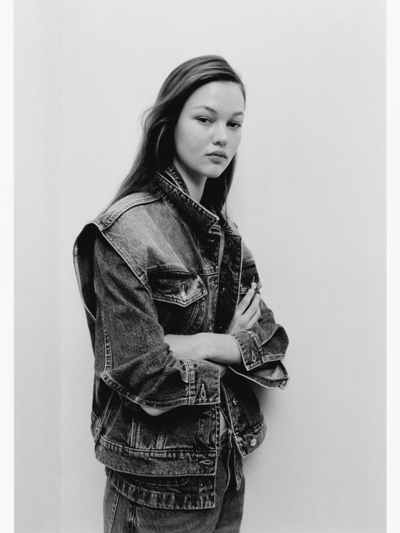 Model wearing a denim vest and jeans standing against a plain white studio background in a black and white portrait.