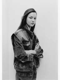 Model wearing a denim vest and jeans standing against a plain white studio background in a black and white portrait.