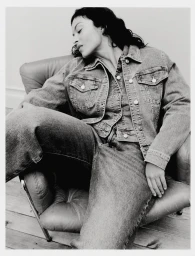 Model seated in an armchair wearing a denim jacket and jeans against a plain background