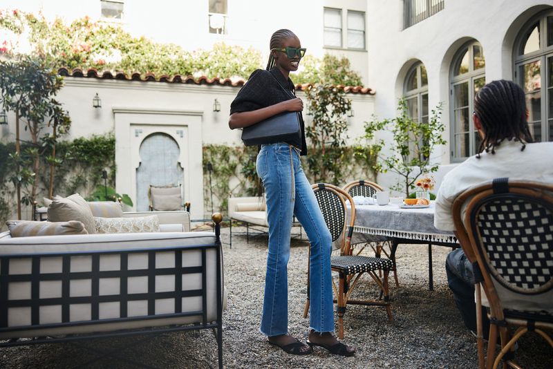 Woman in stylish blue jeans, black jacket and sunglasses poses in an outdoor courtyard with furniture, a table, and a diner in the background.