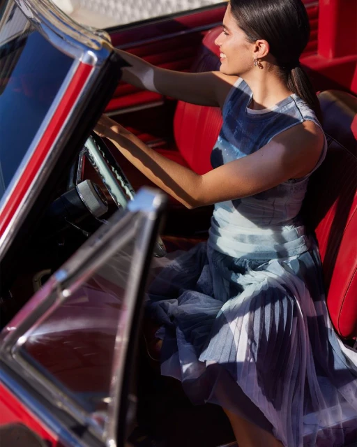 Woman in a flowing dress enters a vintage red convertible car, smiling in bright sunlight. Interior is red leather.