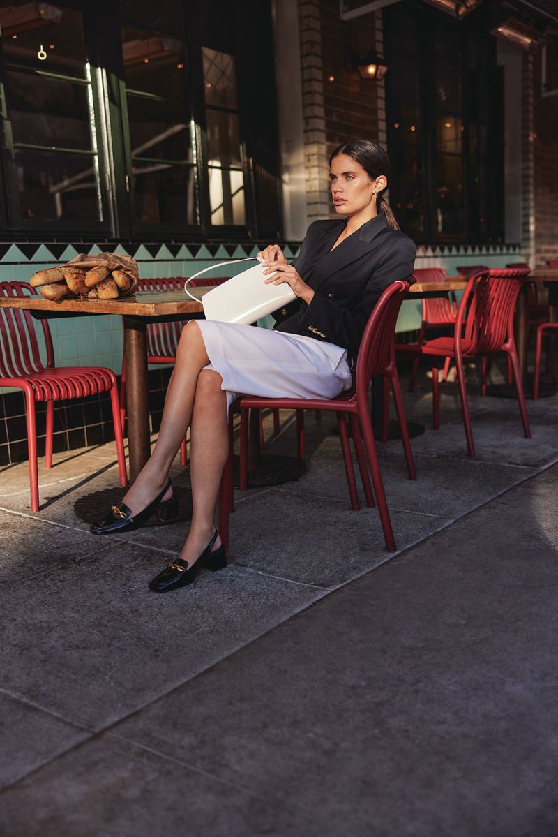 Woman sits at outdoor cafe with white purse and black loafers. She is wearing a dark blazer and light skirt.