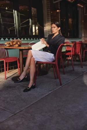 Woman sits at outdoor cafe with white purse and black loafers. She is wearing a dark blazer and light skirt.
