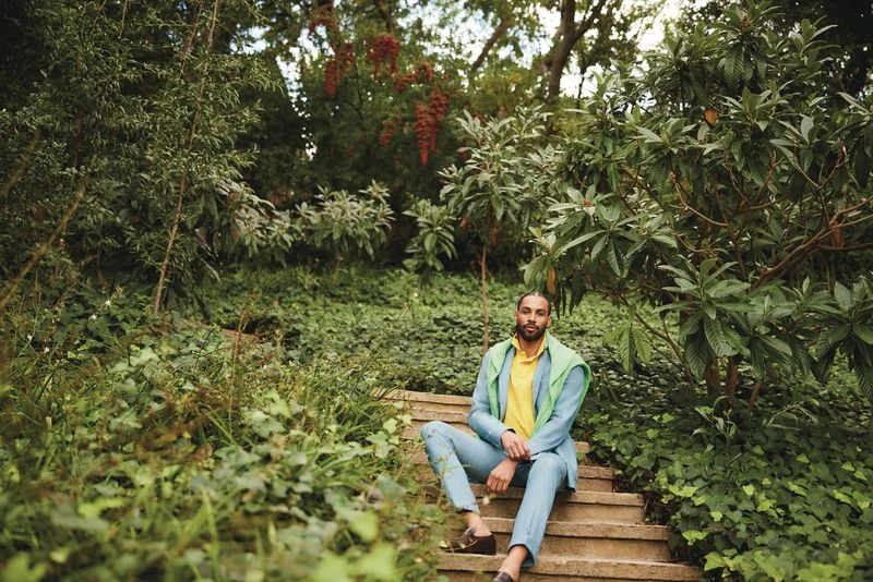 Portrait of adult male in a light blue suit sitting on stone steps surrounded by lush greenery in natural light outdoors.