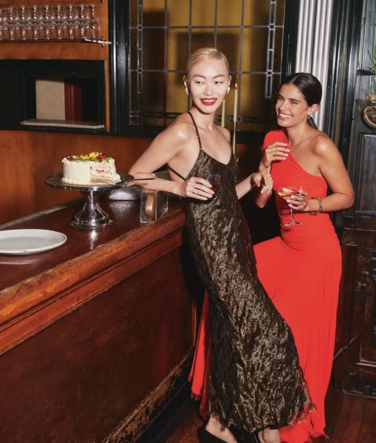 Two women in evening gowns at a bar with cake and cocktails, one fair-skinned, one tan-skinned, dressed for a party or special event.