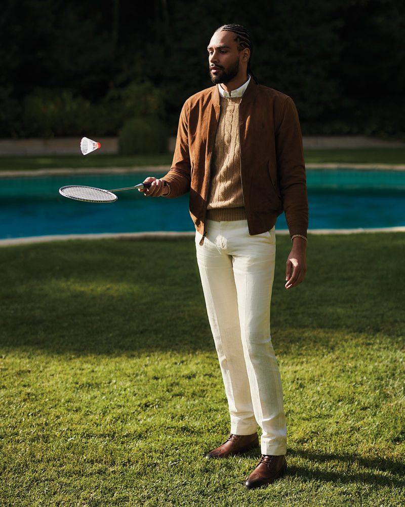 Adult man plays badminton outdoors on sunny day in fashion editorial. He wears a brown jacket, cream pants, and leather shoes.