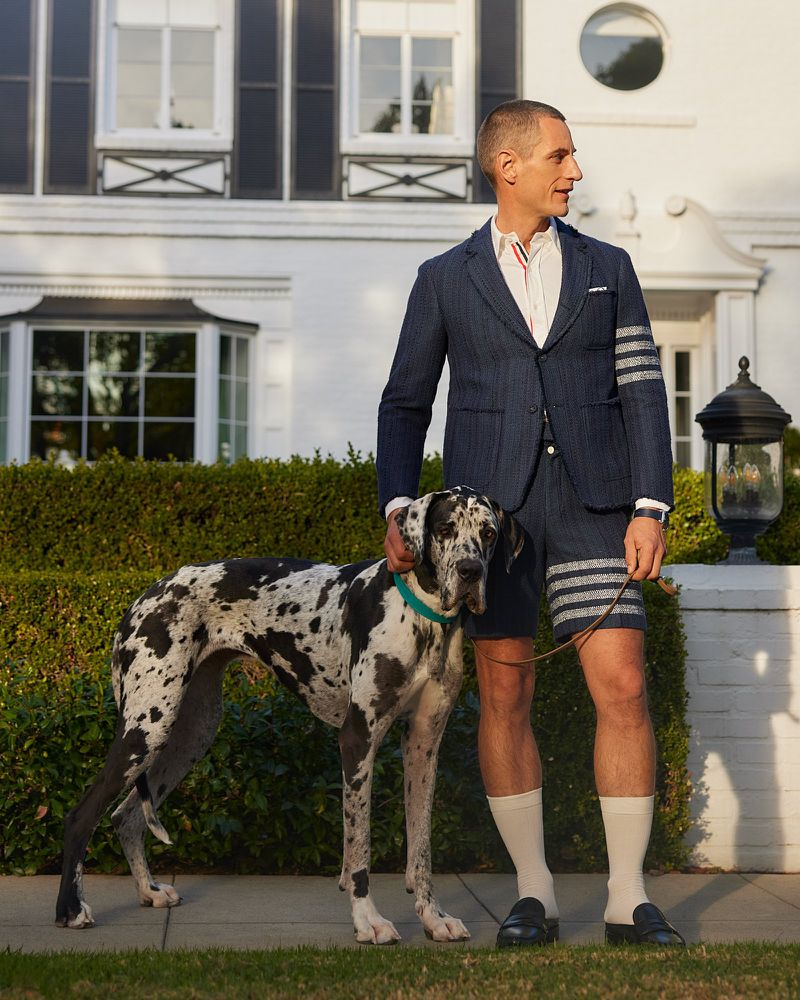 Adult male poses with a Great Dane on a lawn outside a white house with black shutters, showcasing fashion and a pet dog.