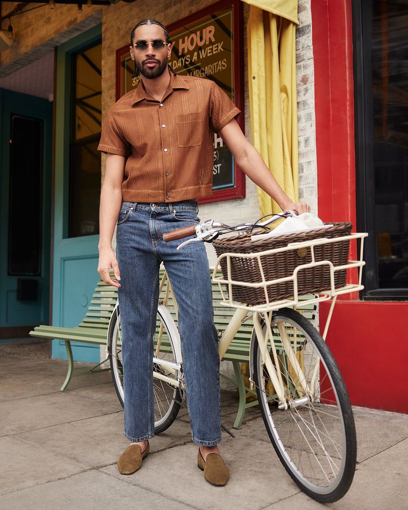 A stylish man in a brown shirt and jeans poses with a bicycle outside a building with a colorful awning, creating a fashionable outdoor scene.