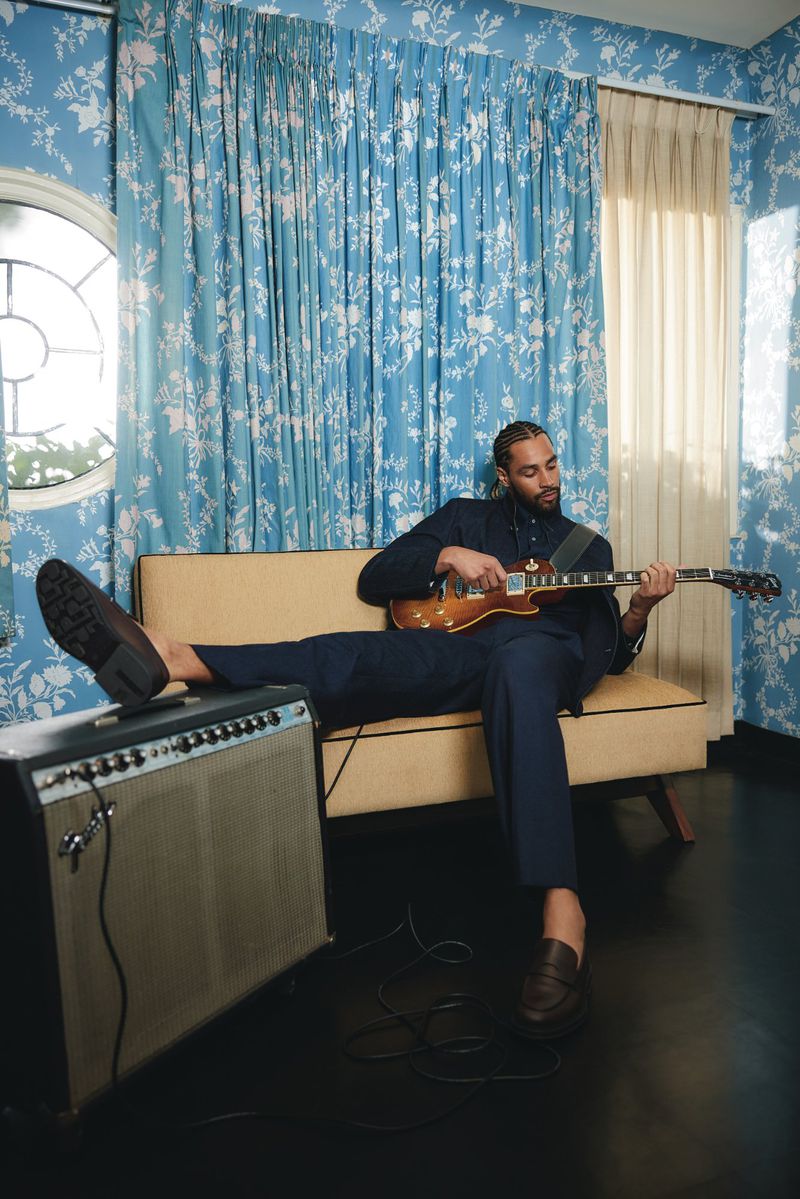 Adult male with an electric guitar relaxing on a yellow sofa with his feet up, next to a speaker in a blue floral room.