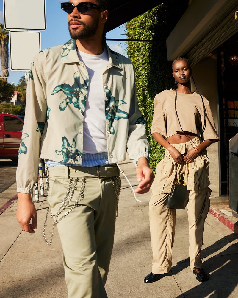 Two fashion models pose on a sidewalk outdoors. An adult male and an adult female wear coordinated beige outfits and sunglasses.
