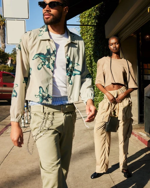 Two fashion models pose on a sidewalk outdoors. An adult male and an adult female wear coordinated beige outfits and sunglasses.