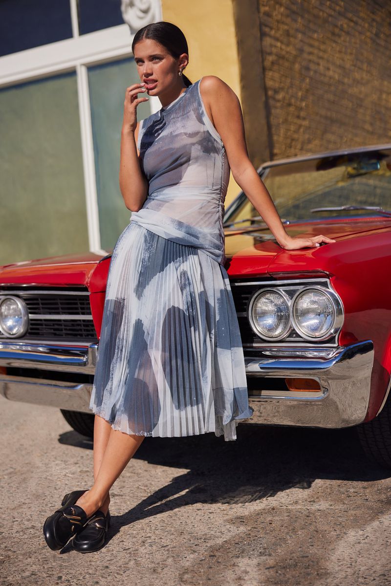 A woman in a sheer dress poses with a red classic car, with sunlight accentuating the scene. She is casually leaning on the hood.