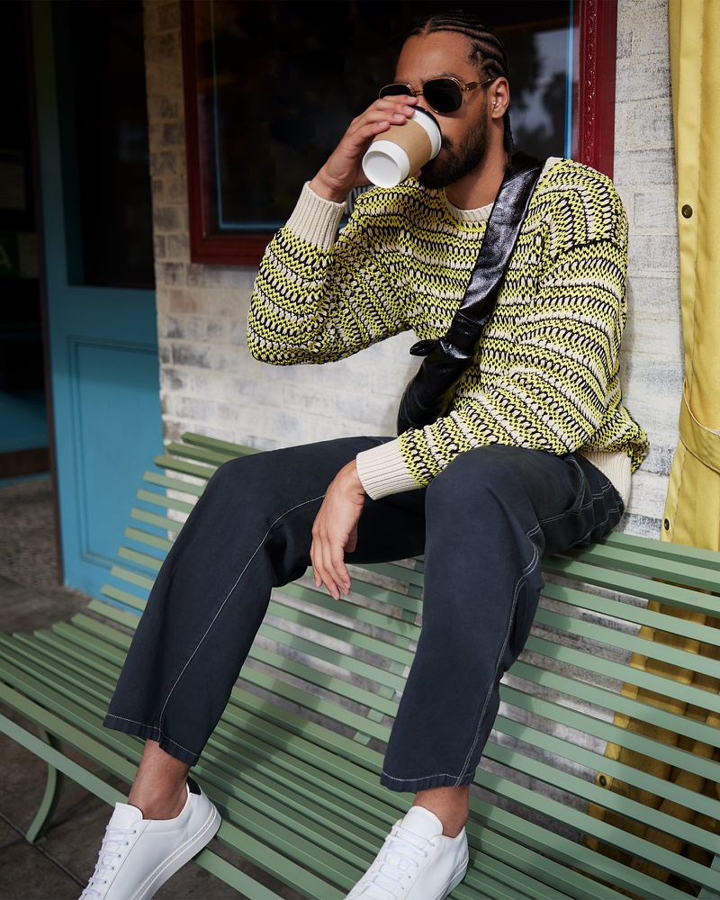 Man wearing a patterned yellow sweater and dark jeans drinks from a paper cup while seated on a green bench outdoors.