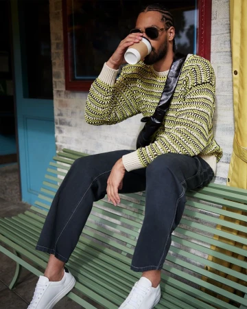 Man wearing a patterned yellow sweater and dark jeans drinks from a paper cup while seated on a green bench outdoors.