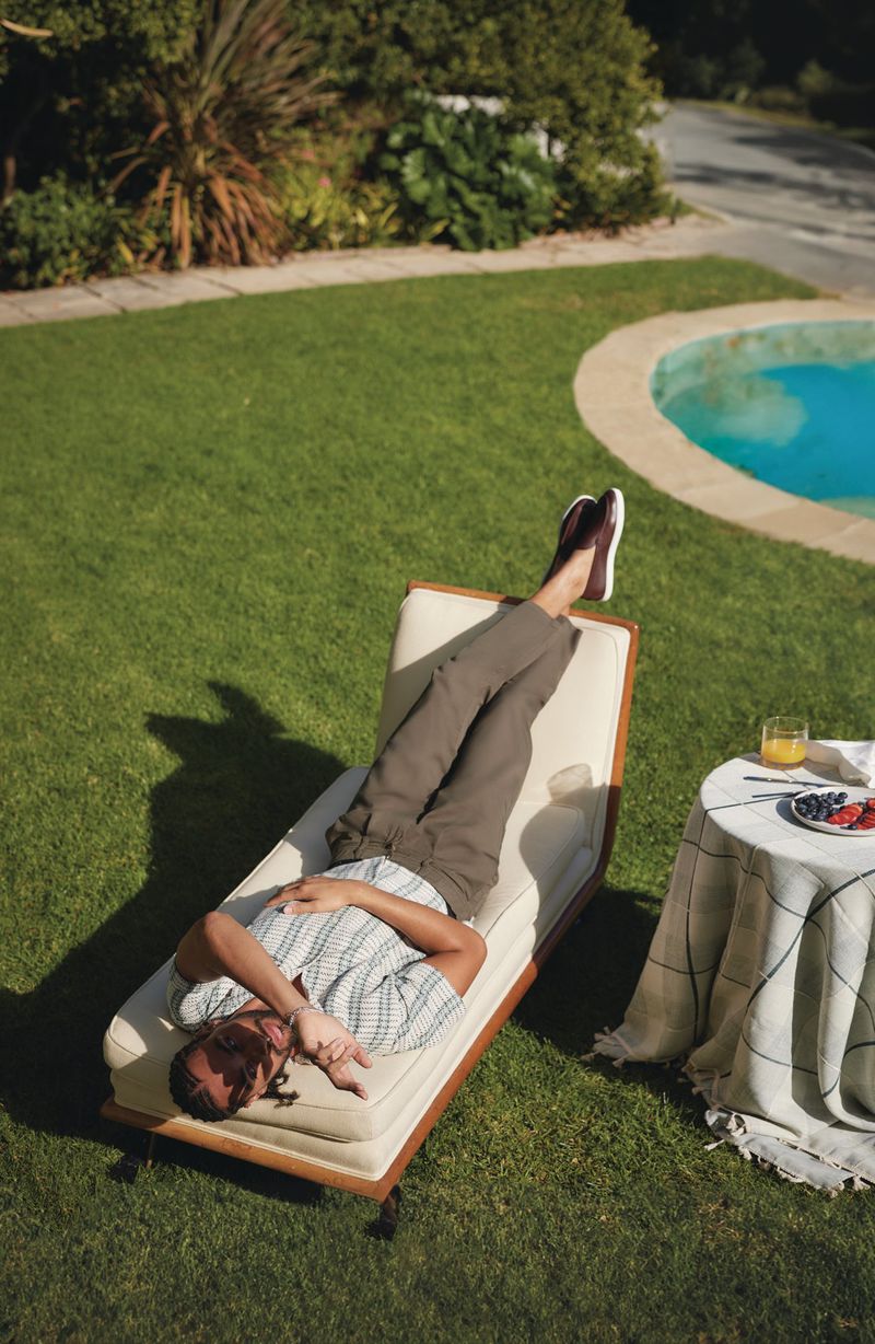 Adult man lies on a lounge chair in a backyard with a small round table topped with fruit and juice. Green grass surrounds him.