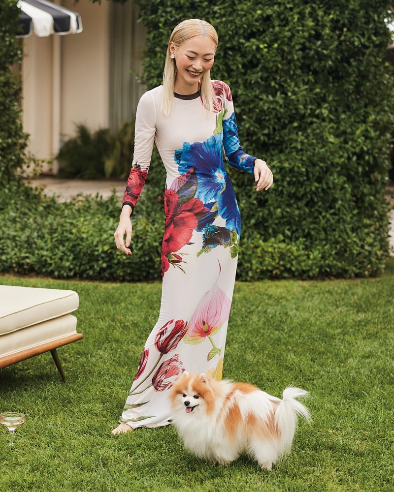 A smiling woman in a floral dress poses on a lawn with her dog. Sunlight illuminates the scene with green hedges in the background.