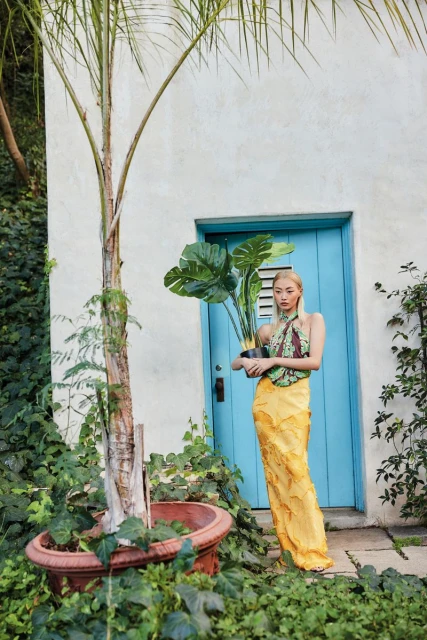Young woman with blonde hair wearing a yellow skirt and halter top, holding a potted plant near a building with a blue door.