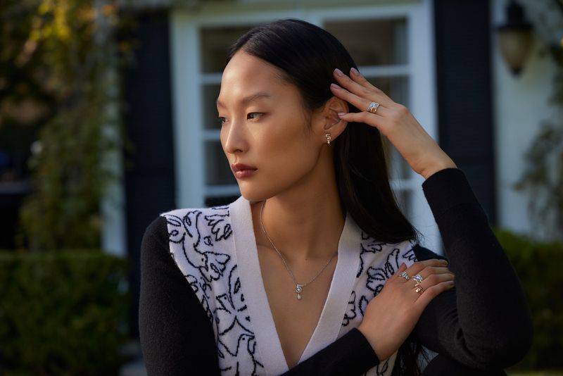 Portrait of a stylish Asian woman wearing jewelry outside near a house, posing with a neutral expression in natural light.