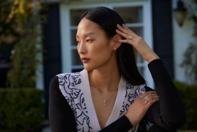 Portrait of a stylish Asian woman wearing jewelry outside near a house, posing with a neutral expression in natural light.