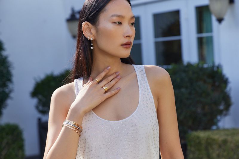 Woman in a sleeveless top wearing gold jewelry including rings, earrings and bracelets outside in front of a white building.