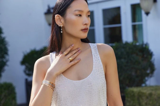 Woman in a sleeveless top wearing gold jewelry including rings, earrings and bracelets outside in front of a white building.