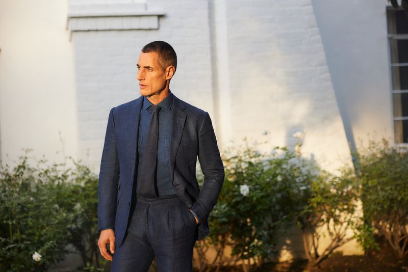 Mature man in blue suit stands outdoors against building with greenery. He is facing left, looking off-camera, and lit by sunlight.