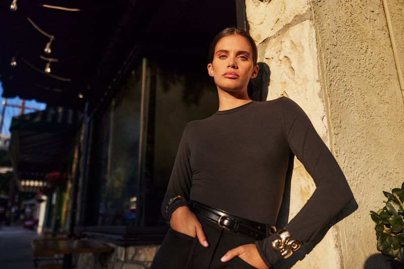 A woman poses outdoors in a black outfit, accessorized with a belt and gold cuffs, leaning against a textured wall with sunlight.