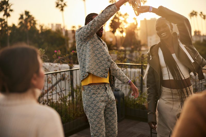 Two friends dance on a rooftop at sunset in designer outfits, palm trees and city skyline background with orange, golden light.