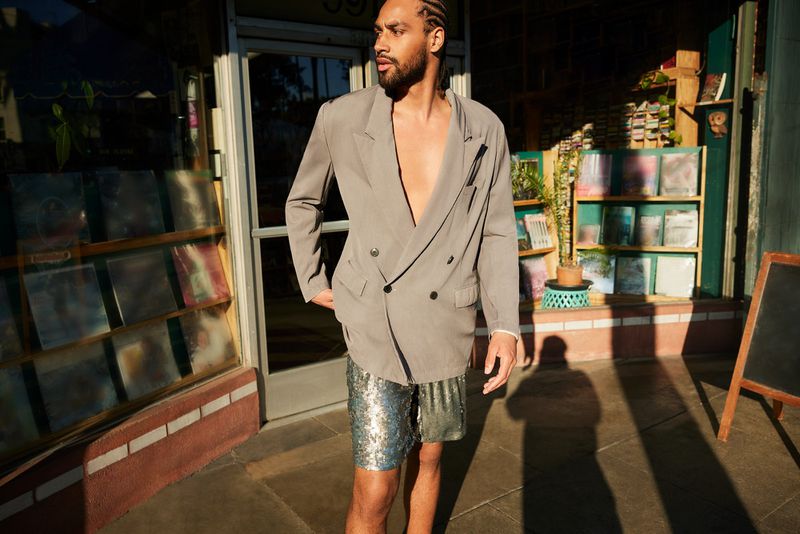 Adult male poses in front of a store in a gray blazer and sequined shorts on a sunny day with sunlight and shadows creating depth.