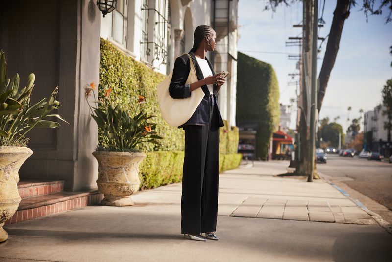 A woman in a dark suit and carrying a tote bag stands on a city sidewalk in the sunlight with trees and buildings in the background.