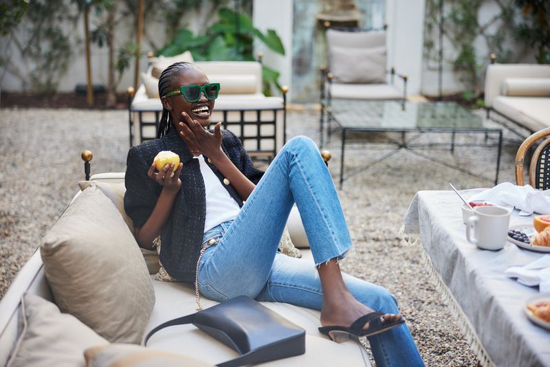 Dark-skinned woman sits outside in jeans, blazer, and sunglasses, holding fruit next to a table with breakfast items and outdoor furniture.