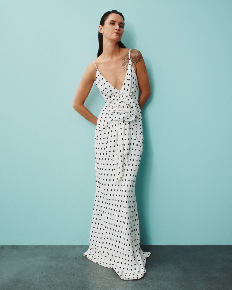 A brunette adult woman poses in a full-length portrait, wearing a sleeveless white gown with black polka dots against a solid cyan backdrop.