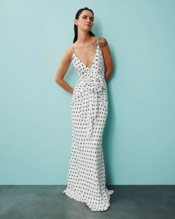 A brunette adult woman poses in a full-length portrait, wearing a sleeveless white gown with black polka dots against a solid cyan backdrop.