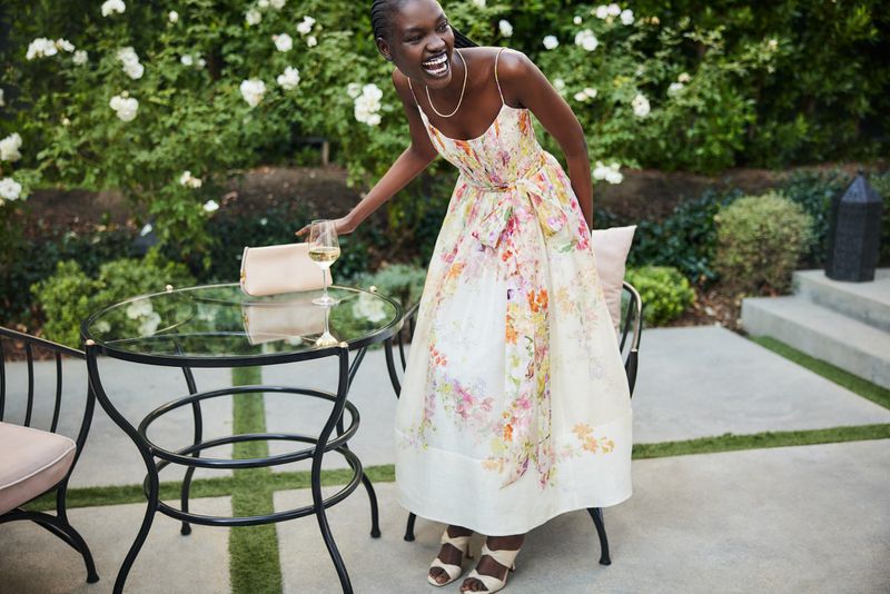 A smiling Black woman in a floral sundress stands near a table with a glass of white wine in an outdoor garden setting.