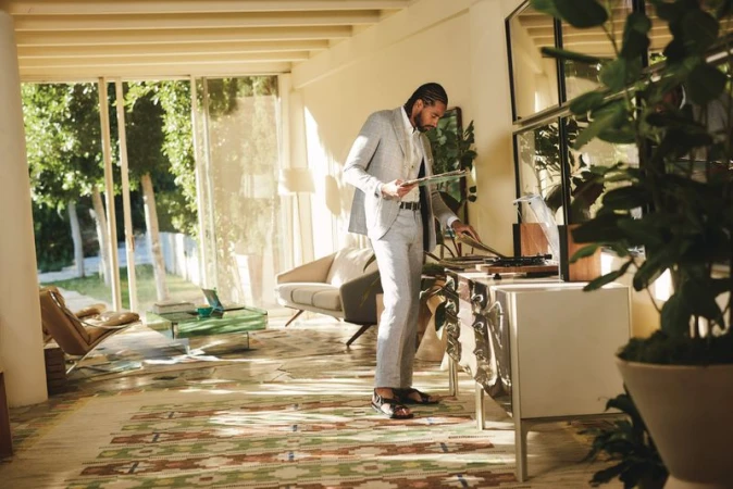 Adult male in a light-grey suit stands in a sunlit living room putting a record on a turntable with indoor plants nearby.