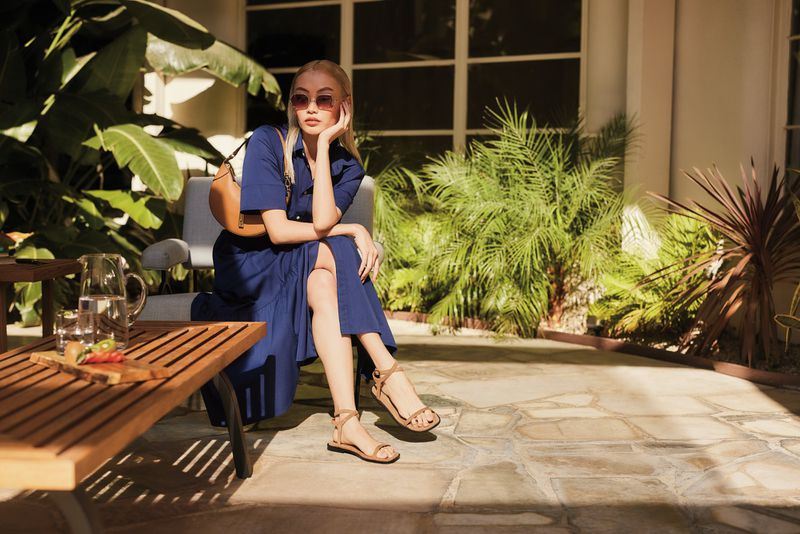Blonde woman in blue dress and sandals sitting on patio furniture with a wood table and glass pitcher, surrounded by greenery.