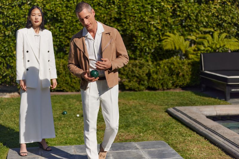 Two people in stylish outfits are playing bocce ball on a lush green lawn with manicured hedges and a modern outdoor lounge set.