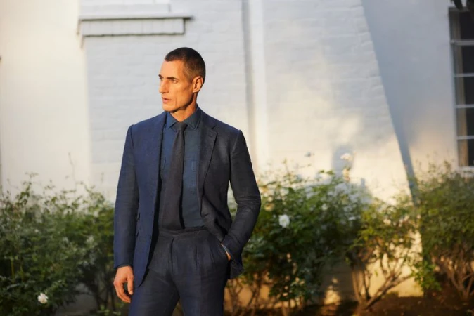 Man in a navy suit and tie standing outdoors against a white building exterior with flowering bushes for Saks SP24