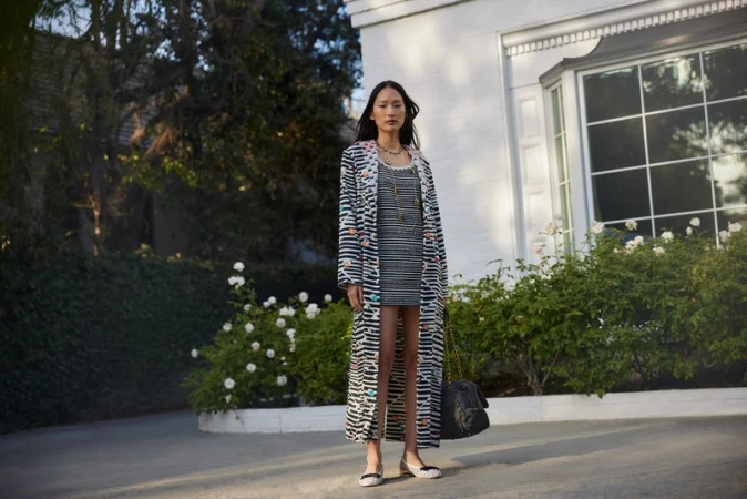 Model standing outdoors wearing a striped coat and dress in front of a white building exterior