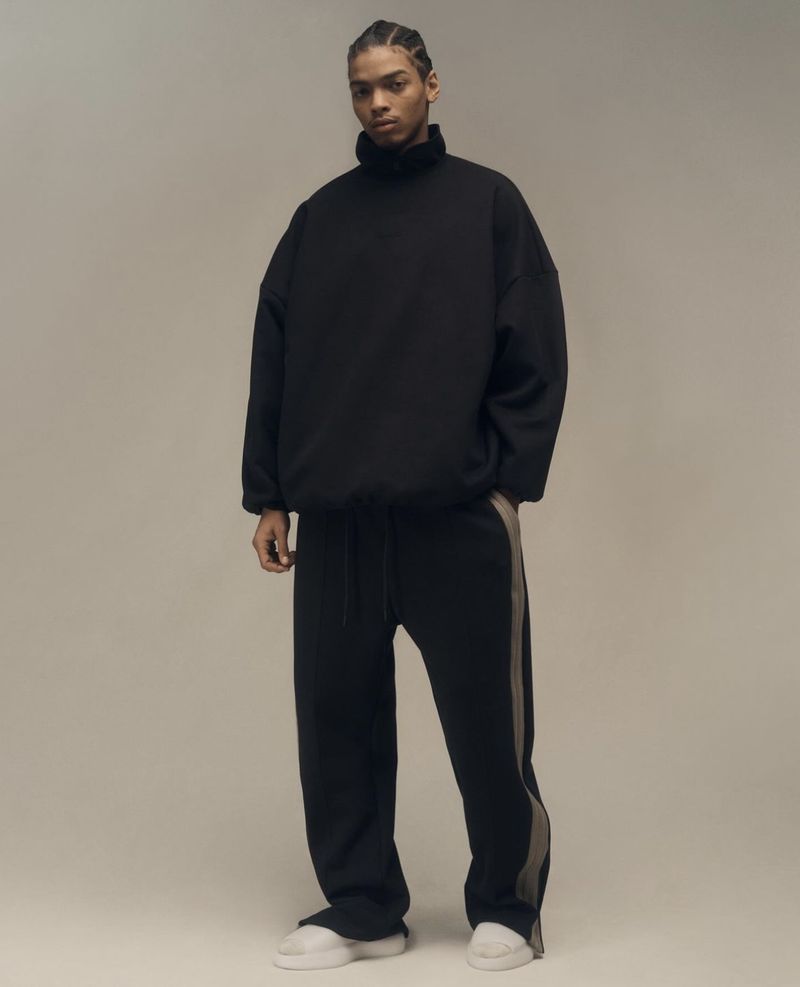 Studio shot of a Black male model in a black turtleneck and matching track pants with gray stripes, posing against a neutral backdrop.