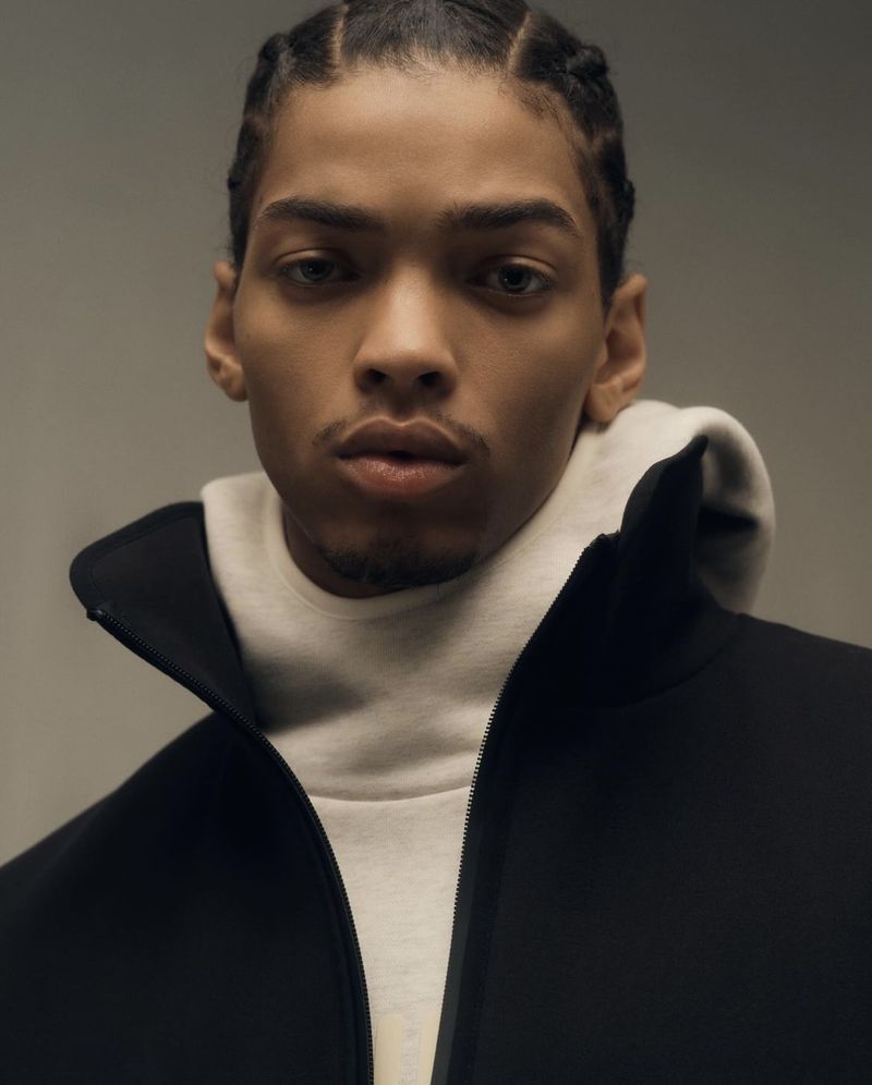Close-up portrait of a young adult male with braided hair, wearing a black zip-up jacket and light grey hooded sweatshirt.