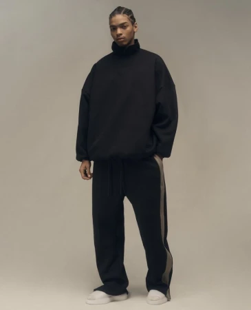 Model wearing a black mock neck sweatshirt and black track pants against a neutral grey studio background
