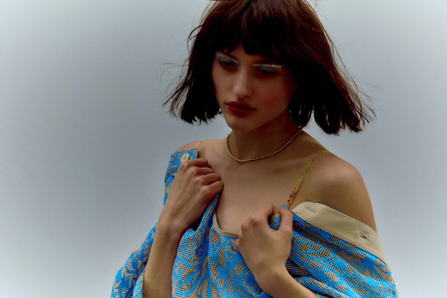 Close-up of a woman with dark hair wearing a blue patterned garment and a gold necklace against a neutral background.