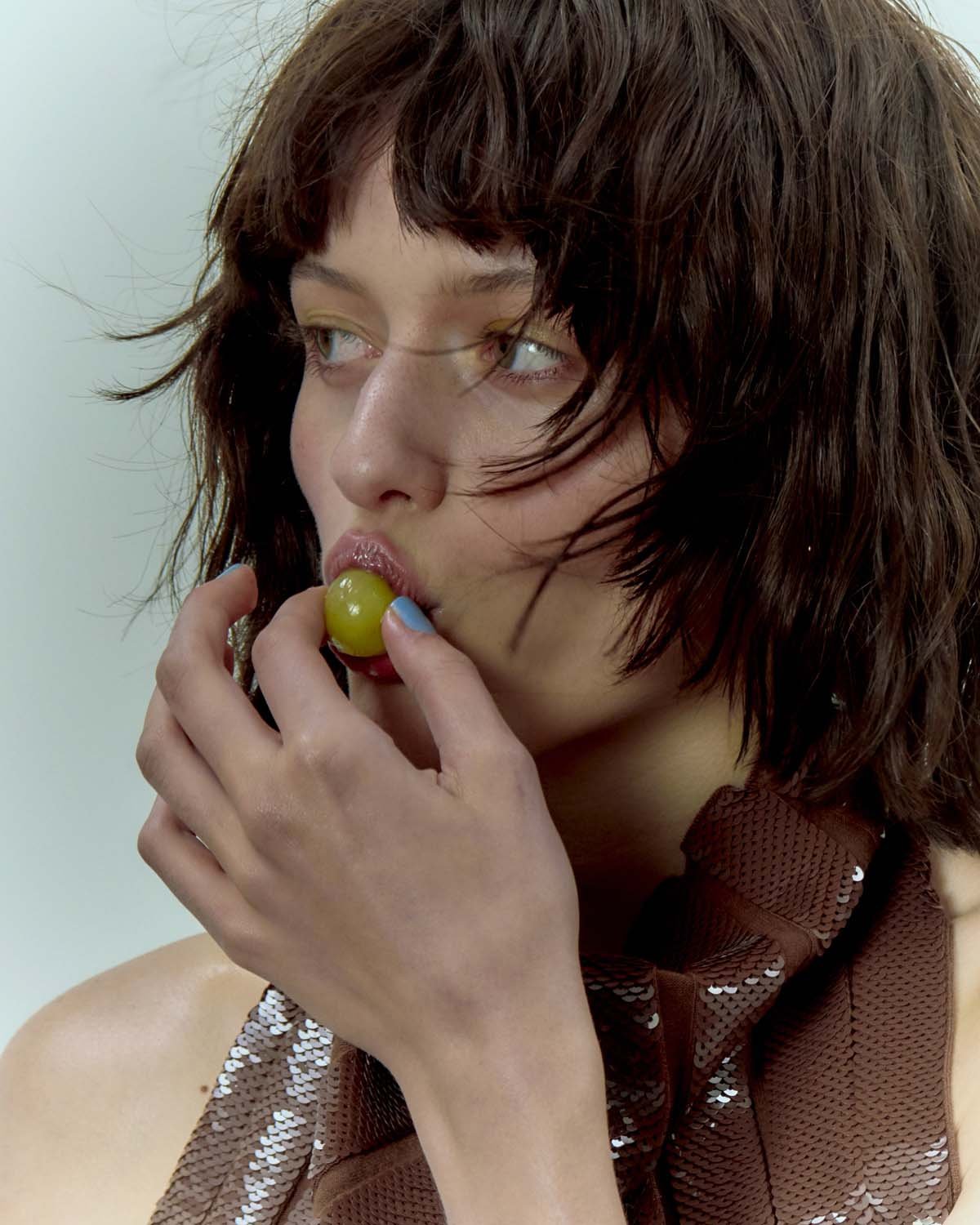 A woman with short brown hair and sequined top has a green grape in her mouth in this eye-level portrait in a white studio setting.