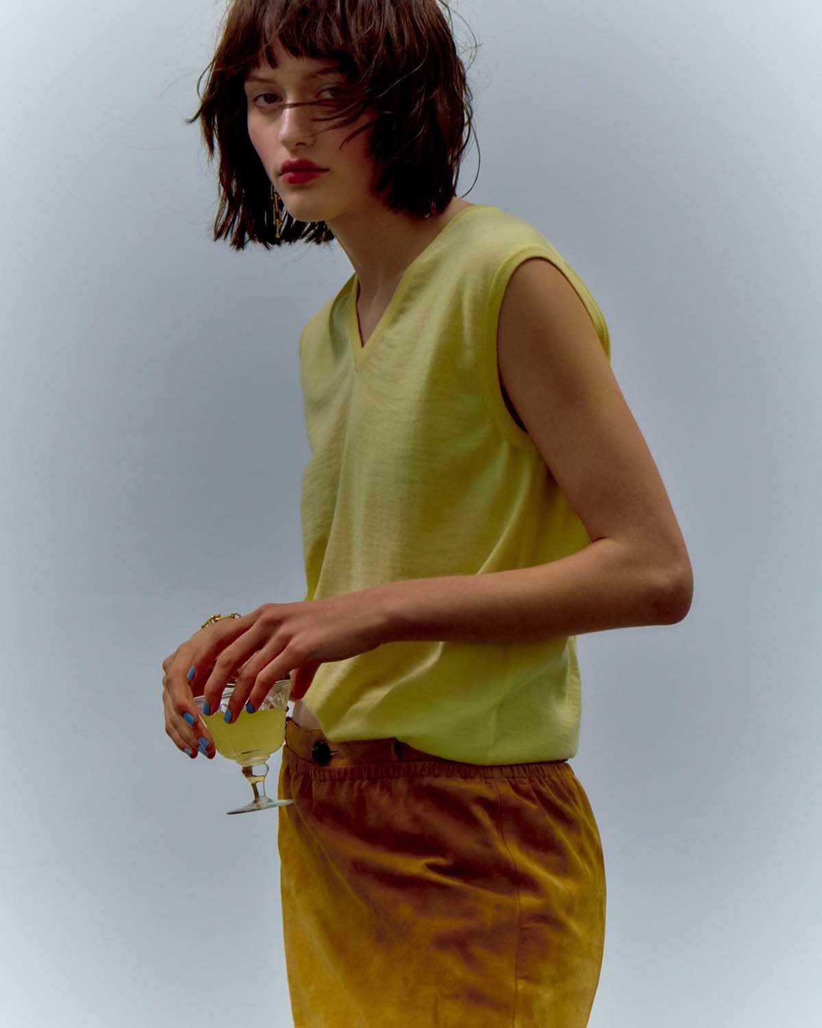Young woman with bobbed hair in a chartreuse top and mustard skirt, holding a glass of liquid against a bright, neutral background.
