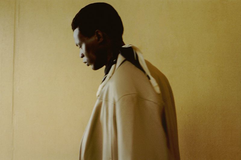 Profile of an adult male model in a beige jacket against a light yellow backdrop, showcasing contemporary fashion and understated style.