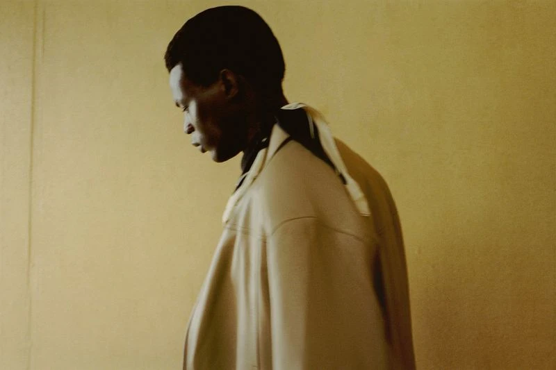Profile of an adult male model in a beige jacket against a light yellow backdrop, showcasing contemporary fashion and understated style.