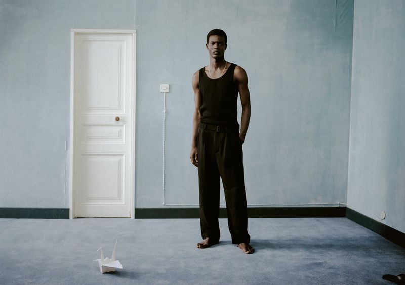Adult male in black tank top and pants stands in a light blue room with a white door and paper crane on the grey floor.